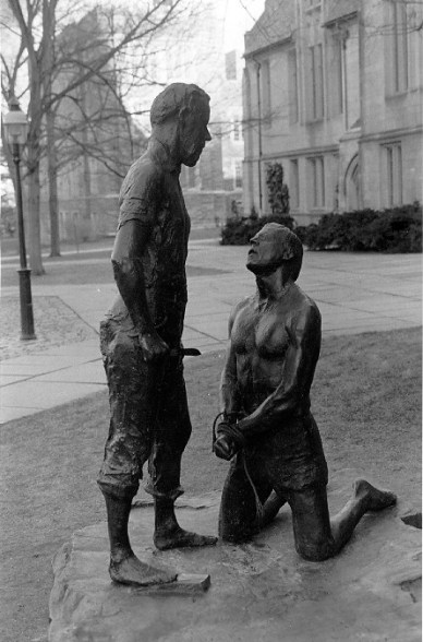 A sculpture of a man standing over another man kneeling on the ground. The kneeling man's hands are bount in front of him and he is looking up imploringly.