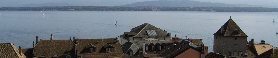 A photo of mountains in the distance, with Lake Geneva in the middle ground and rooftops of Nyon in the foreground.