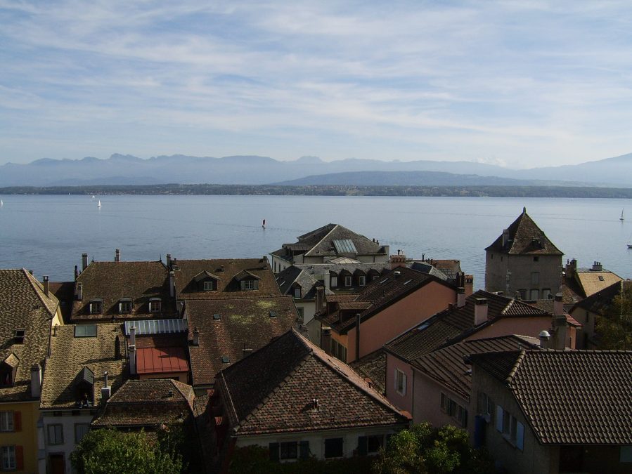 A photo of mountains in the distance, with Lake Geneva in the middle ground and rooftops of Nyon in the foreground.