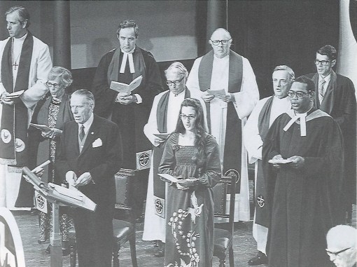 Black and white photo from 1977 of eight men and two women in clerical garb with Uniting Church Scarfs, holding service booklets and singing.