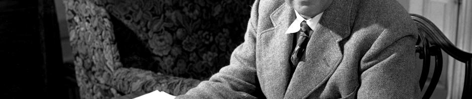 Black and white photo of a middle-aged, balding white man sitting at a desk looking towards the camera while holding papers in his hands.