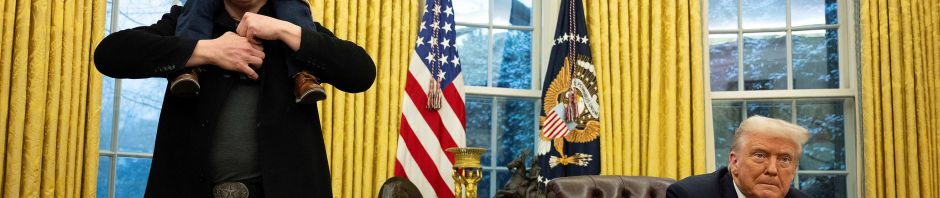 Photo of Elon Musk in a black coat and cap with his son on his shoulders standing in the Oval Office while President Donald J. Trump sits at the Resolute Desk.